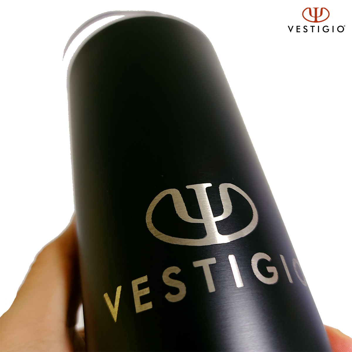Mano sostiene termo negro con logo y texto VESTIGIO, fondo blanco.