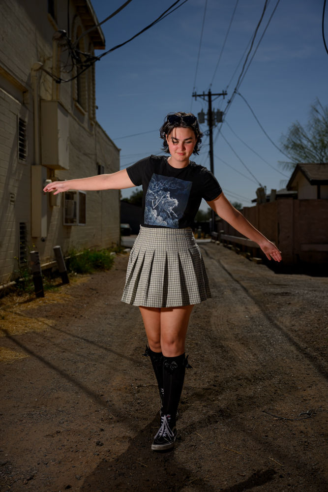 Young person in black shirt and plaid skirt balances in a sunlit alley. Overhead wires and a blue sky set the urban scene, evoking focus.