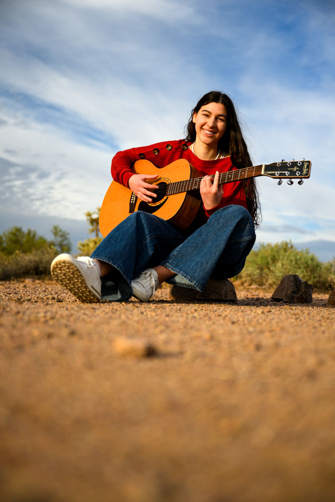 Smiling person in a red sweater and jeans plays acoustic guitar outdoors on a sunny day, sitting on a rocky terrain with a blue sky background.