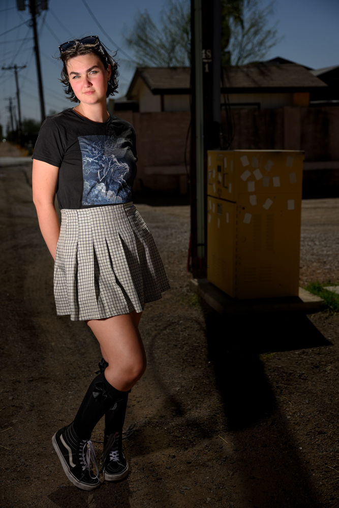 A girl stands in a dirt alley wearing a graphic tee, plaid skirt, and knee-high socks. Sunlit background with power lines and buildings.