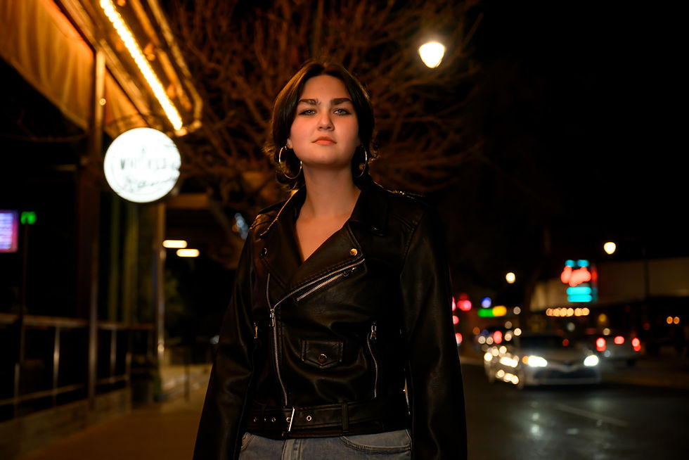 A woman in a black leather jacket stands on a city street at night, with bright streetlights and blurred car lights in the background.