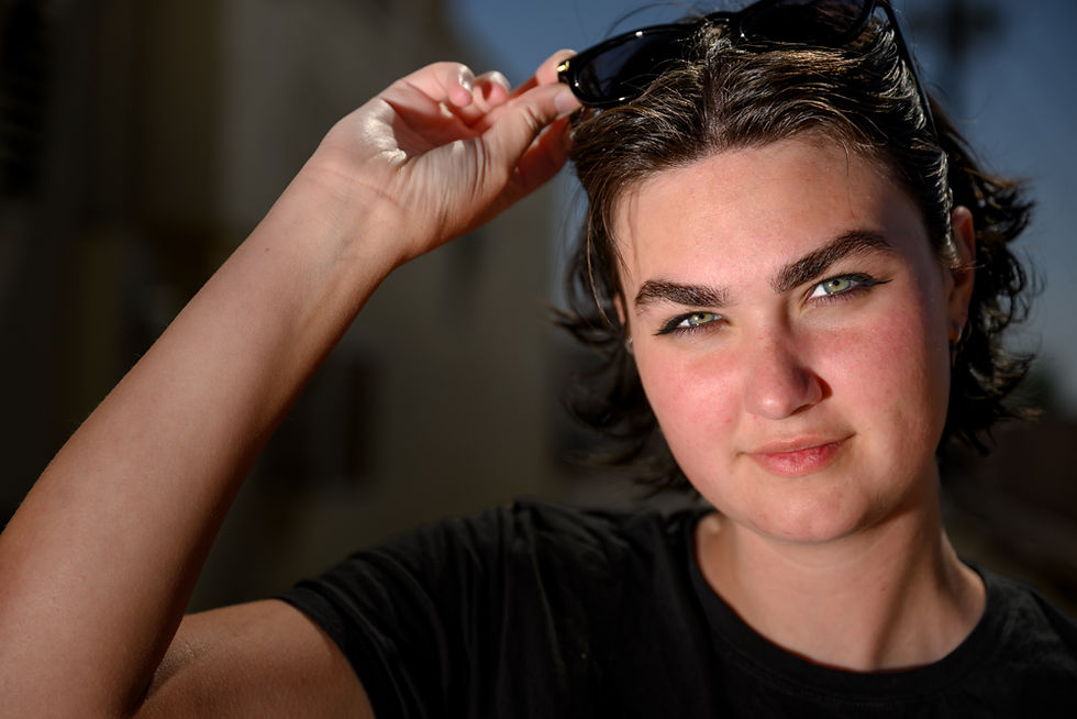 Person with short dark hair adjusts sunglasses, outdoors. Sunlit face with slight smile. Wearing a black shirt against a blurred background.