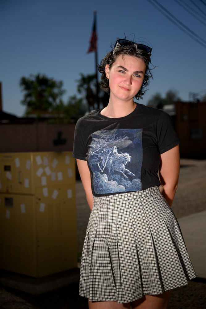 Woman in a black graphic tee and plaid skirt stands outdoors, smiling. Background shows a flag and trees. Sunlight creates a warm ambiance.