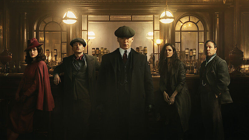 Five people in vintage attire stand in a dimly lit bar with ornate decor. They look serious, wearing hats and coats, evoking a moody atmosphere.