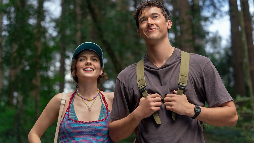Smiling man and woman in casual attire hike in a forest, wearing backpacks. Sunlight filters through trees, creating a cheerful atmosphere.