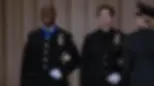 Two uniformed officers stand smiling on stage, one with a blue medal ribbon. They're in a formal setting with beige curtains in the backdrop.