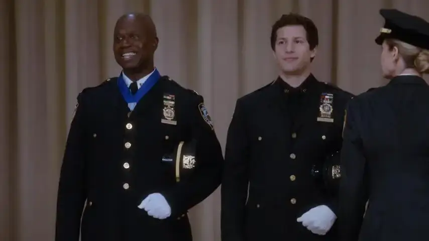Two uniformed officers stand smiling on stage, one with a blue medal ribbon. They're in a formal setting with beige curtains in the backdrop.