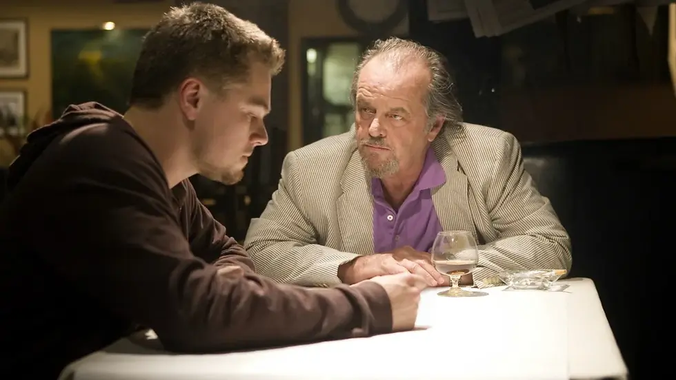 Two men lean over a table in a dim restaurant, one in a blazer and purple shirt, sharing a tense conversation.