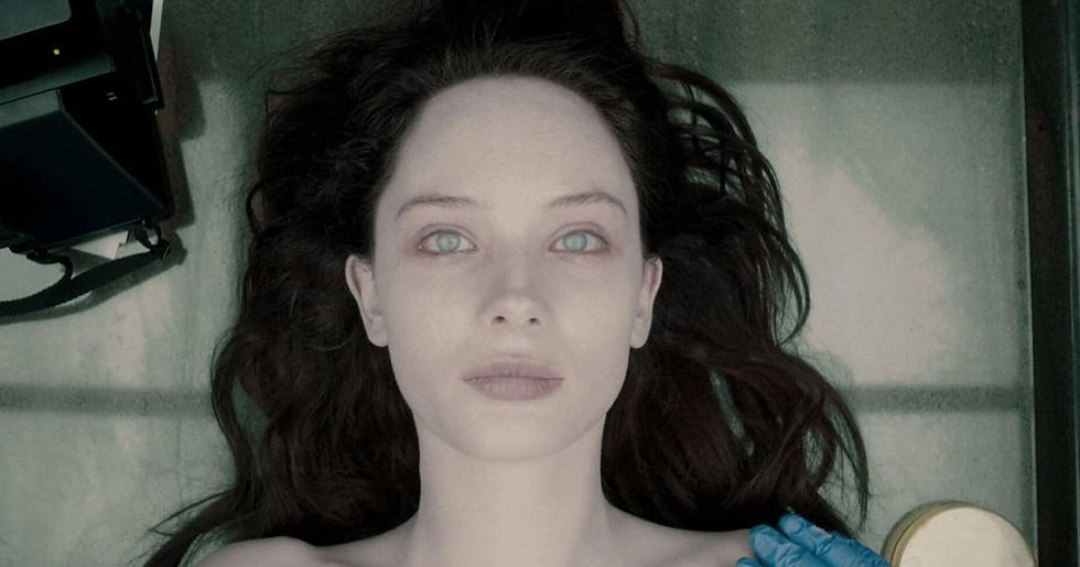 Pale woman lies on a metal table, dark hair spread out, as a blue-gloved hand touches her shoulder in a sterile room.