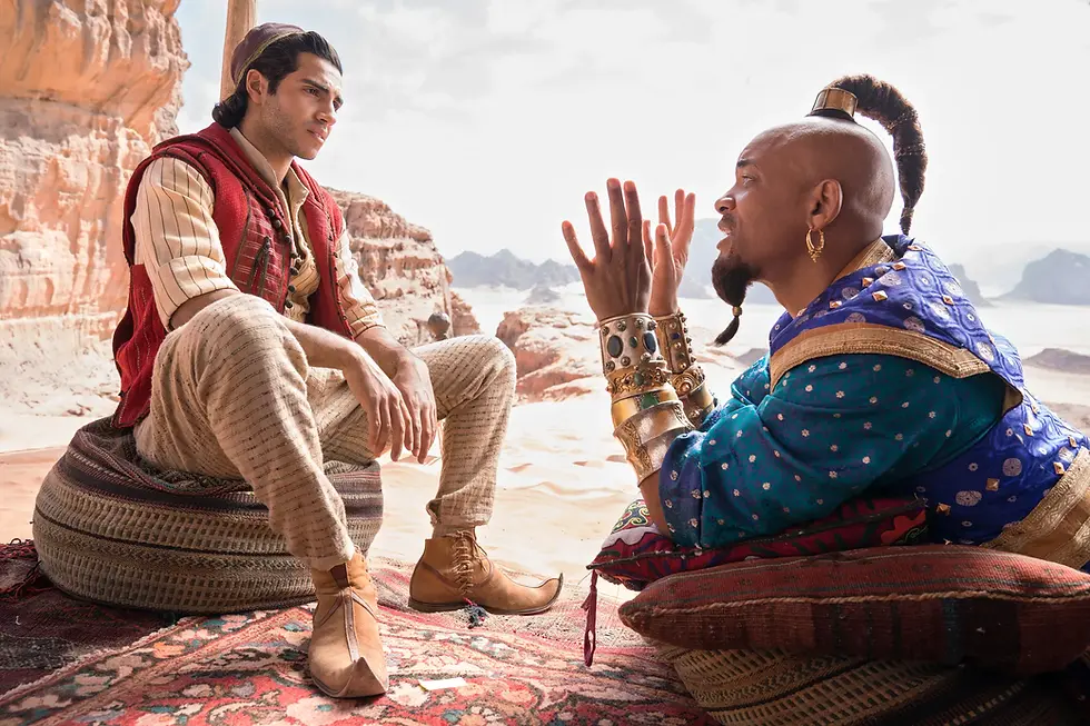 Two men in colorful desert robes sit on cushions in a canyon, talking face to face as one gestures animatedly.