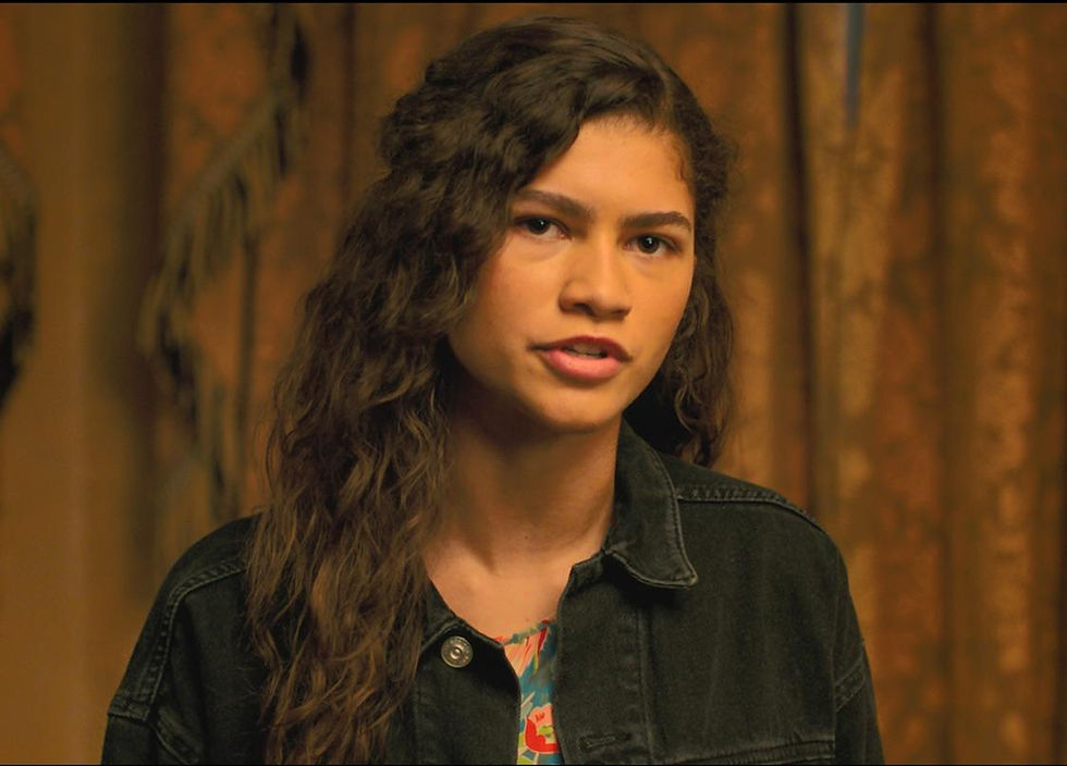 Young woman with curly hair and denim jacket speaks against a brown patterned curtain. She appears focused and expressive.