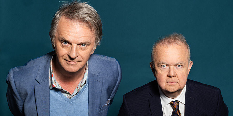 Two stern-looking men in suits pose against a teal studio backdrop, one leaning forward, both facing the camera.