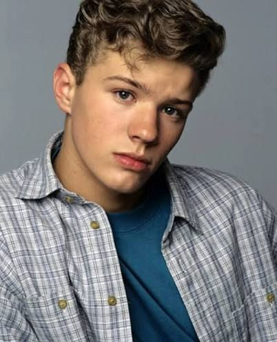 Portrait of a young man in a plaid shirt over a blue tee, posing against a gray background with a neutral expression.