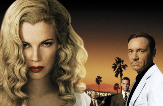 Close-up of a blonde woman with a serious stare, beside men in suits and a man in a white coat, with Hollywood sign at sunset.
