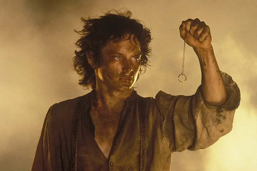Dusty man in a warm smoky scene holds up a small ring pendant on a chain, staring intently to the side.
