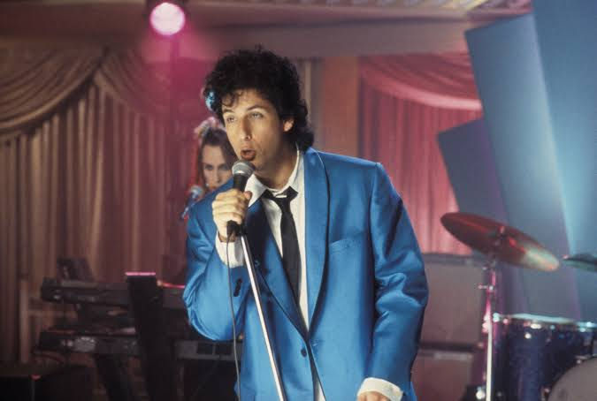 Singer in a bright blue suit sings into a microphone on a pink-lit stage, with band instruments and curtains behind him.