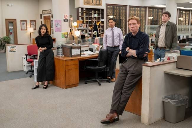 Four serious coworkers stand in a newsroom office with desks and glass partitions, one leaning on a counter.