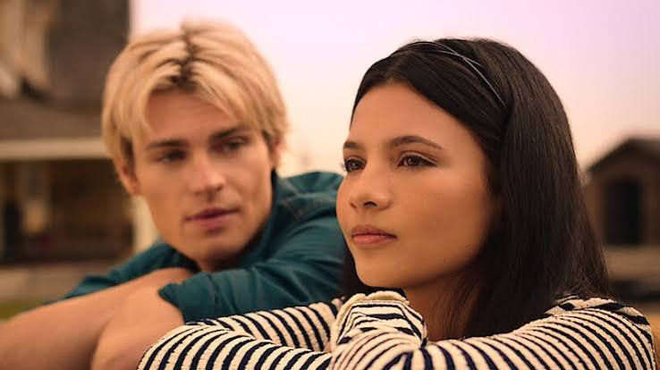 Two young adults lean on their arms outdoors, a blonde man watching a dark-haired woman in a striped top with a thoughtful mood.