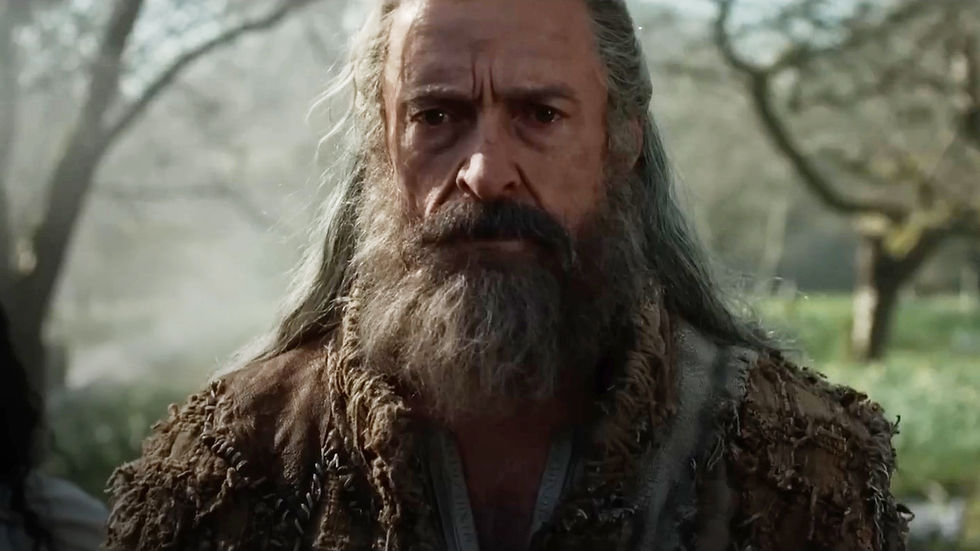 Bearded man with long hair and worn clothing stands outdoors, surrounded by blurred, green foliage. Serious expression, rustic setting.