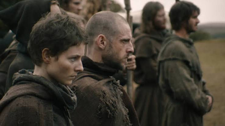 Somber medieval-clad group stands outdoors, with a short-haired woman and bald man in front, looking ahead.