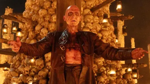 Bald man in a dark robe stands with arms outstretched before a wall of skulls and candles, creating a sinister, eerie scene.