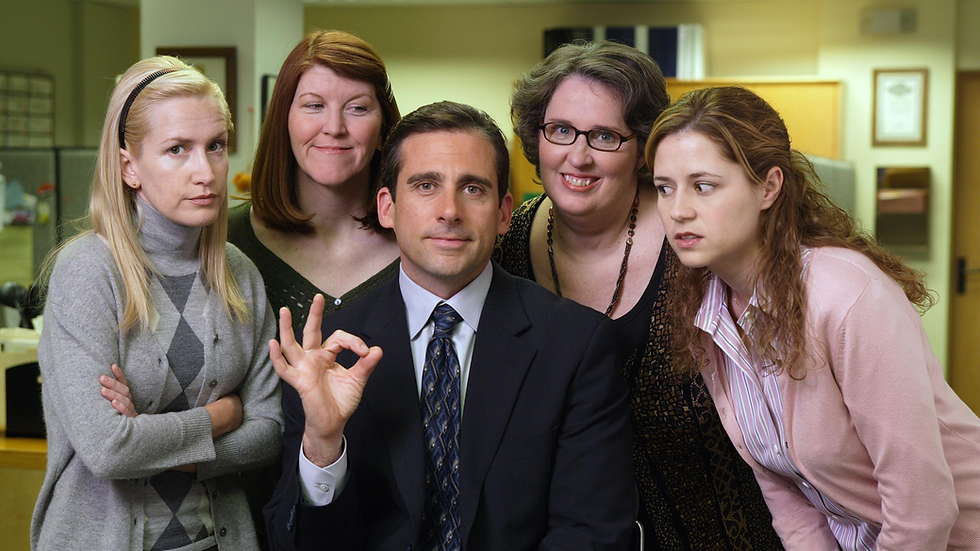 Five office workers pose in a warm-lit office; a suited man makes an OK sign while the women crowd around him, smiling and skeptical.
