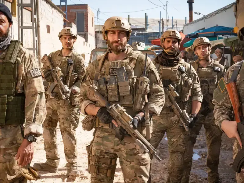 Armed soldiers in camouflage and helmets stand in a dusty street, facing camera with serious expressions; ISAF patch visible.