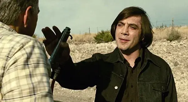 Man in black jacket points a handgun at another man in a barren desert, tense standoff under a pale sky.