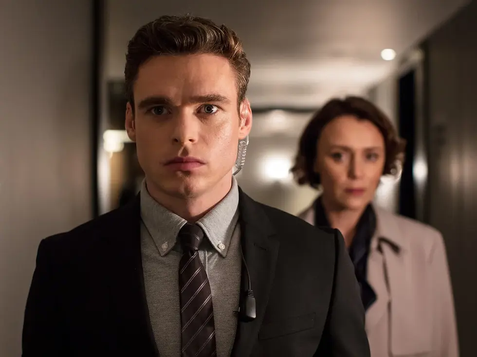 Serious man in suit with earpiece stands in a hallway, with a blurred woman behind him, tense and focused.