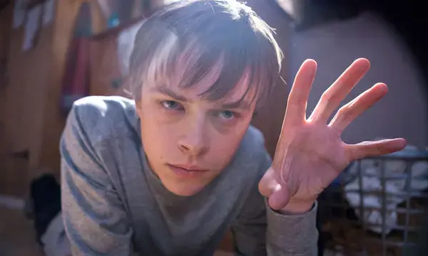 Boy in a gray shirt reaches toward the camera outdoors, with an intense stare and sunlit background.