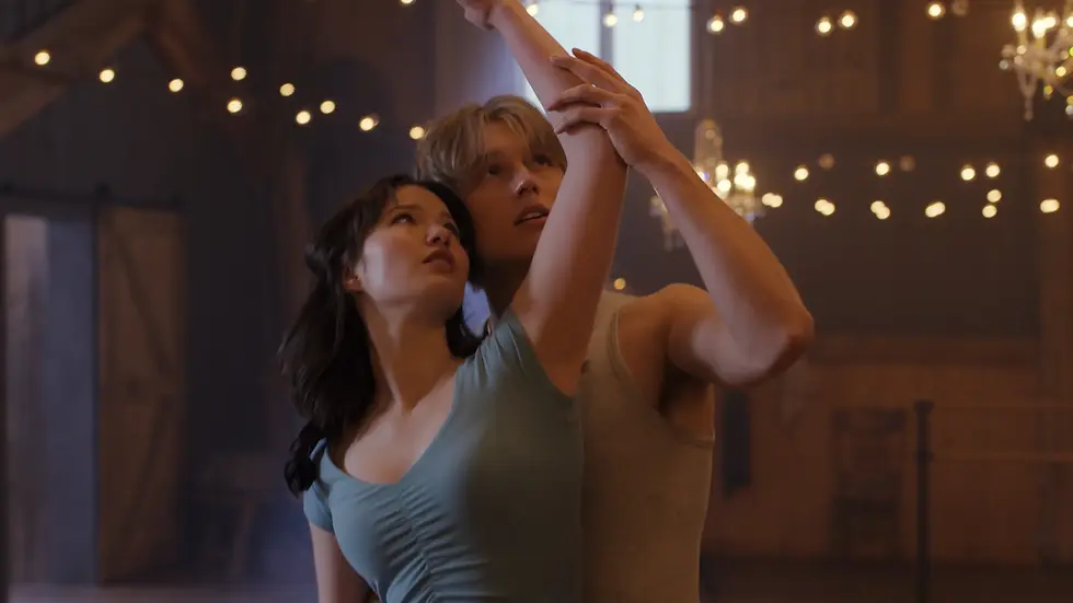 Couple dancing closely in a warmly lit wooden hall with string lights and chandeliers, raising joined arms in a tender pose.