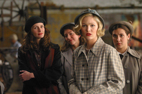 Four women in vintage coats and hats stand indoors, looking serious in a rustic, dimly lit setting.