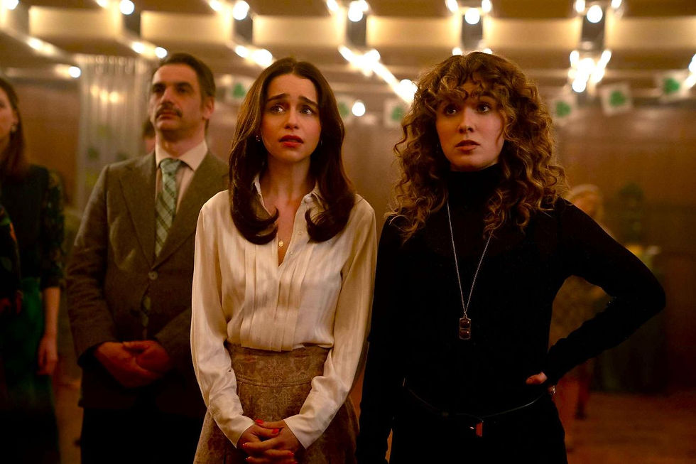 Three tense adults stand in a warm-lit elevator lobby, two women foregrounded, one man behind, all looking worried and alert.