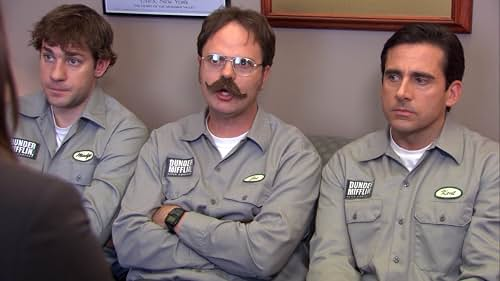 Three Dunder Mifflin men in gray shirts sit on a couch in an office, looking serious and attentive.