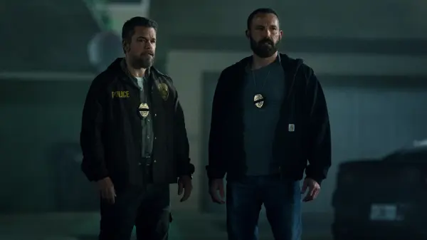 Two men with badges stand in a dimly lit room, appearing serious. One wears a jacket labeled "POLICE". The setting is somber and tense.