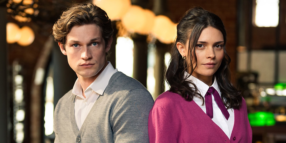 Two people in a warmly lit room, one in a gray sweater and the other in a pink cardigan. Both look serious. Blurred lights in background.