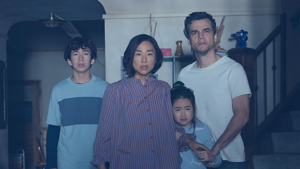 An anxious family of four stands together in a dim home hallway, looking toward something offscreen.