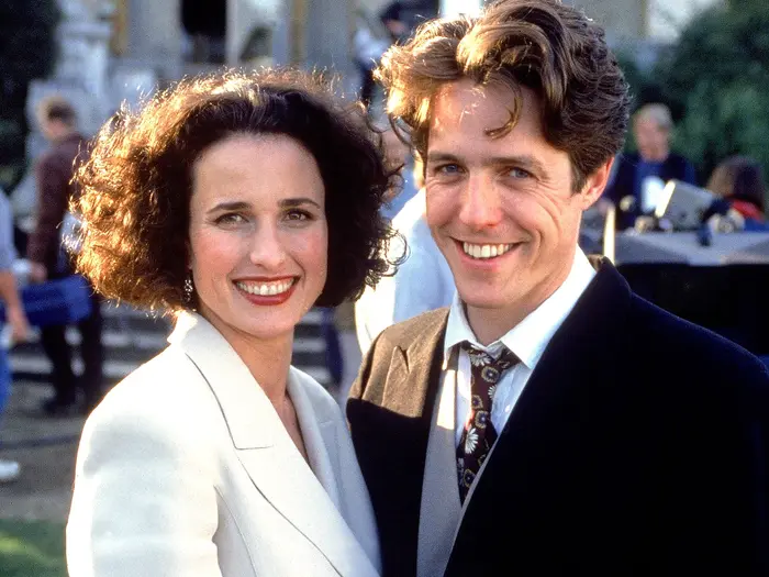 Smiling man and woman in formal clothes pose outdoors at a sunny event, with blurred people and buildings in the background.