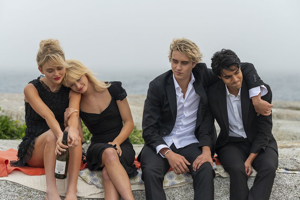 Four people in black attire sit on a seaside stone wall. Two women comfort each other, one holding a wine bottle. Calm, overcast mood.
