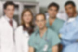 Five doctors in white coats, teal scrubs, and a blue gown pose in a hospital hallway, looking serious.