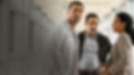 Three serious adults stand in a bright hospital corridor, a doctor in white coat beside a man and woman holding a phone.