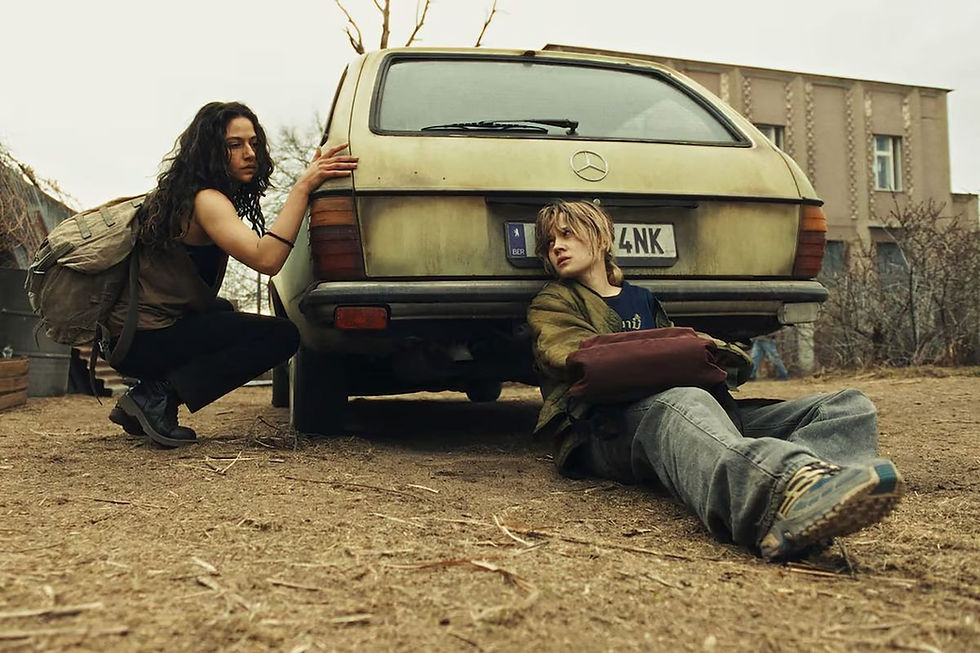 Two young people crouch and sit by a dusty beige Mercedes van in a barren yard, looking tense and wary.