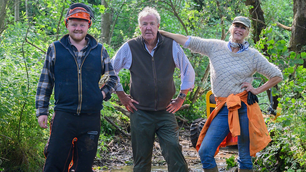 Three smiling people pose in a leafy woodland; one wears a chainsaw helmet, with work clothes and an orange jacket tied at waist.