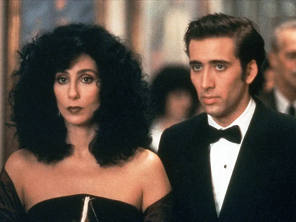 Woman with big black hair in a strapless black gown stands beside a man in a tuxedo at a formal event.
