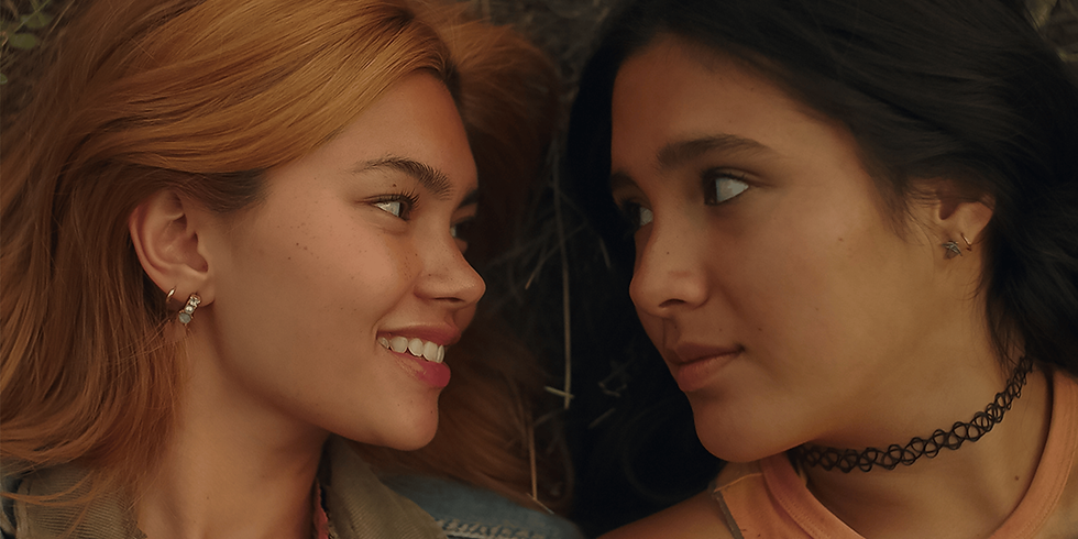 Two women lying on grass, facing each other with smiles. One has blonde hair, the other dark. Warm tones create a gentle and intimate mood.