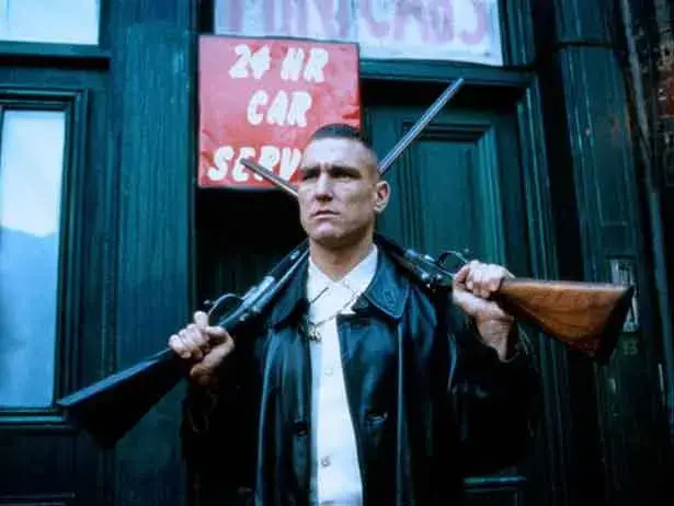 Serious man in a black leather jacket carries two rifles over his shoulders outside a dark building with a 24 HR CAR SERV sign.