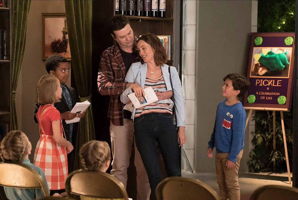 A scene from Single Parents shows two adults standing together as children watch during a school event.