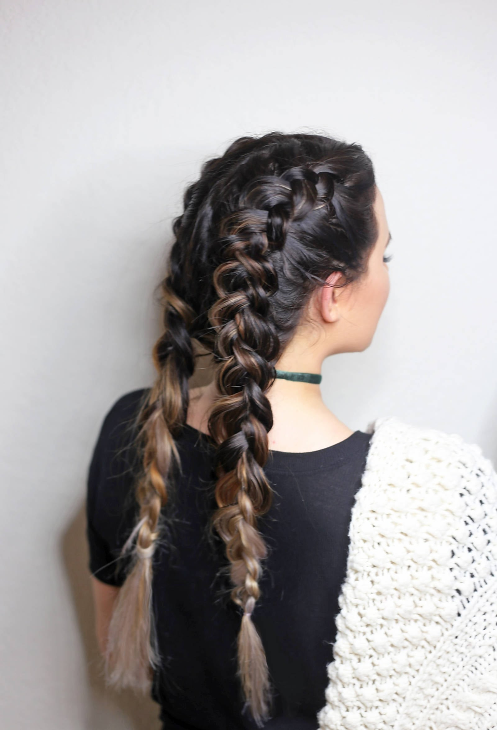 Back view of a woman with two long braids, black top and cream knit shoulder, against a plain white background.
