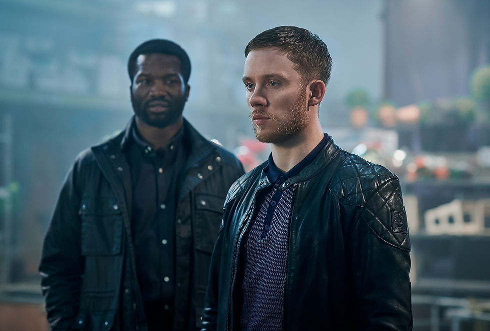Two serious men in black leather jackets stand in a hazy industrial space, one in front and one blurred behind.