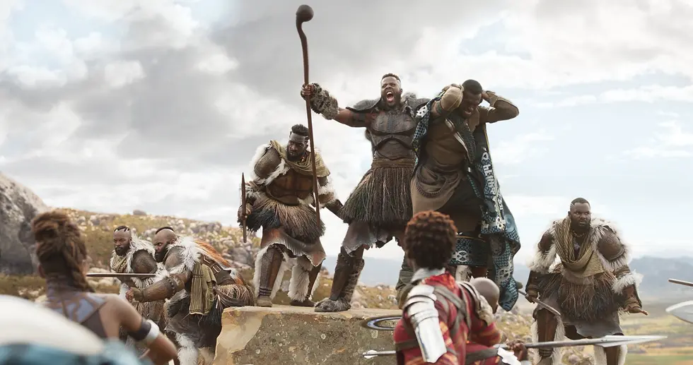 African warriors battle on a rocky hillside, one shouting with a staff as others brace with spears under a cloudy sky.